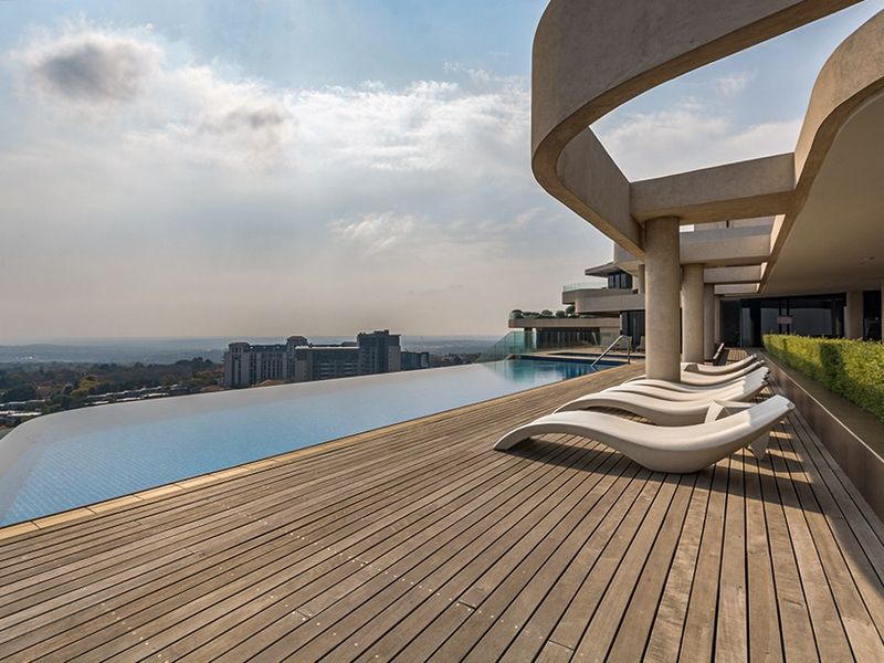 A luxury infinity edge swimming pool featuring timber decking and panoramic city views, set within a modern architectural building structure.