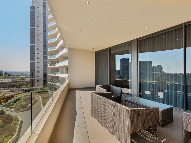 Spacious covered balcony featuring modern tiled flooring, a sleek glass balustrade, and recessed ceiling lighting, offering expansive views of the surrounding urban landscape.