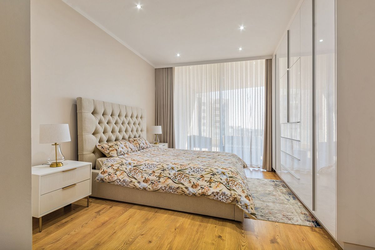 A modern, well-maintained bedroom featuring high-gloss built-in wardrobes, recessed lighting, and large windows with floor-to-ceiling curtains leading to a balcony.