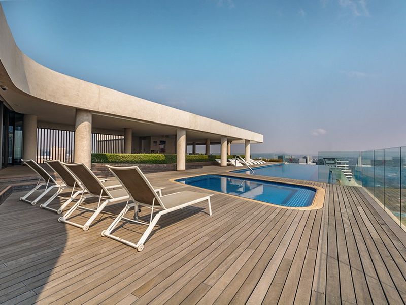 A modern rooftop infinity pool area featuring extensive timber decking, glass balustrades, and a covered terrace with panoramic city views.