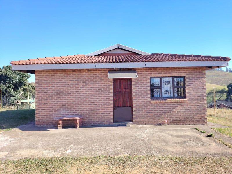 A single-story face brick house featuring a tiled roof, security gate, and a concrete patio area set in a rural landscape.