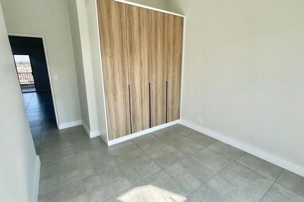 A bright, modern bedroom featuring neutral walls, large-format grey floor tiles, and contemporary wood-finish built-in cupboards.