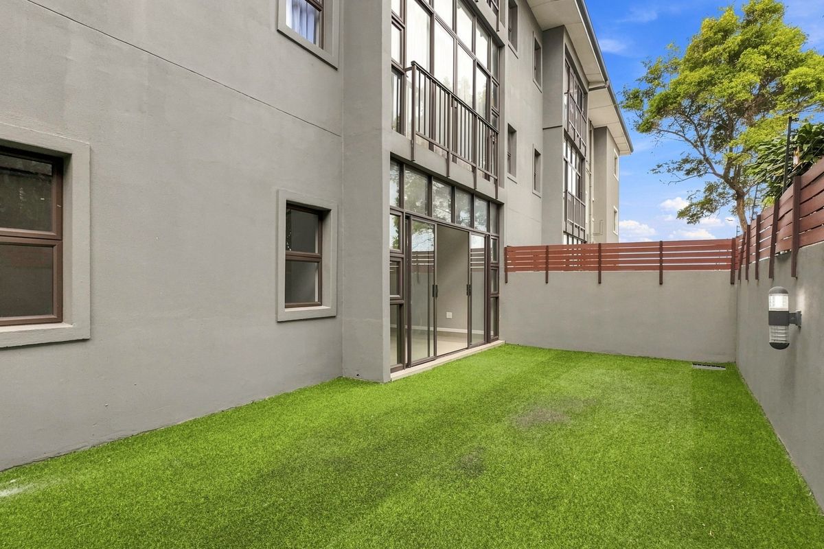A private rear garden area featuring artificial turf, a perimeter fence, and direct access to the apartment via sliding glass doors.