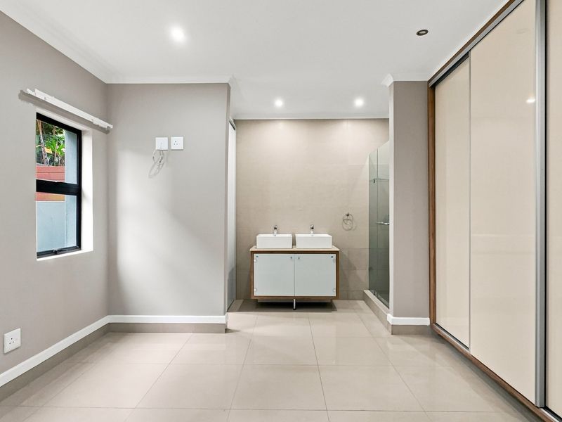 Modern bathroom featuring a double vanity, glass-enclosed shower, and integrated sliding door wardrobe with neutral tiled flooring and recessed lighting.