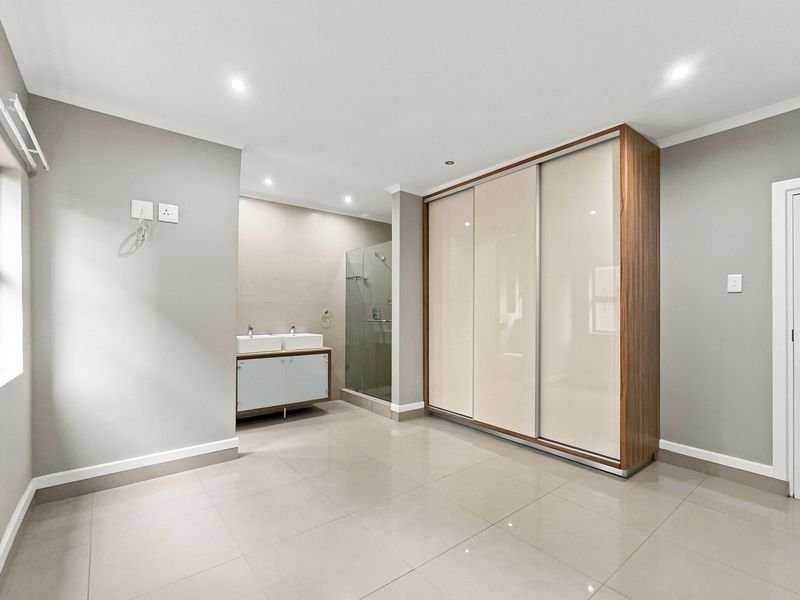 Spacious bedroom featuring a large built-in sliding wardrobe, neutral tiled flooring, and an integrated en-suite bathroom with a double vanity and glass shower.