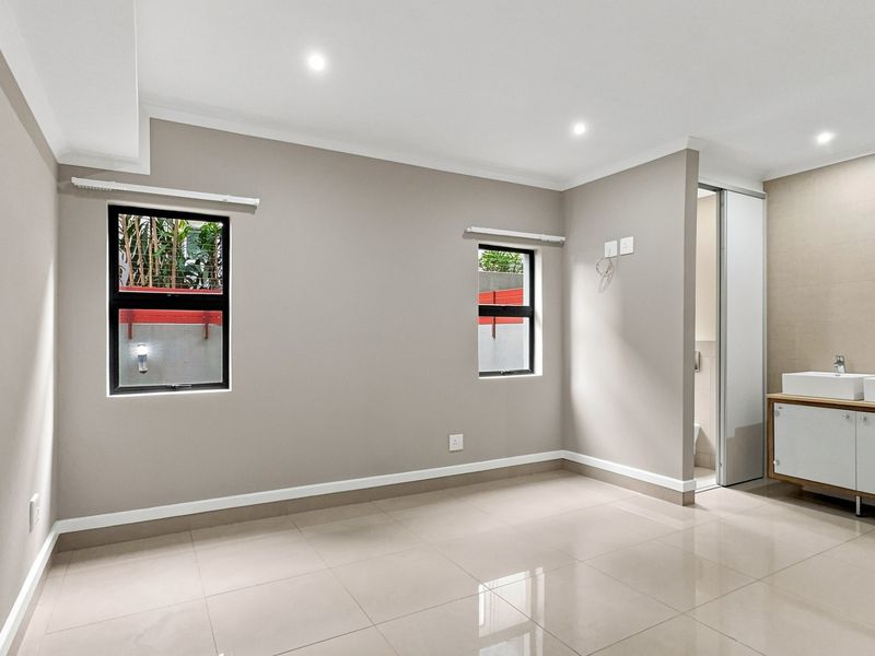 A bright, empty bedroom featuring neutral walls, tiled flooring, recessed lighting, and an en-suite bathroom with a modern double vanity.