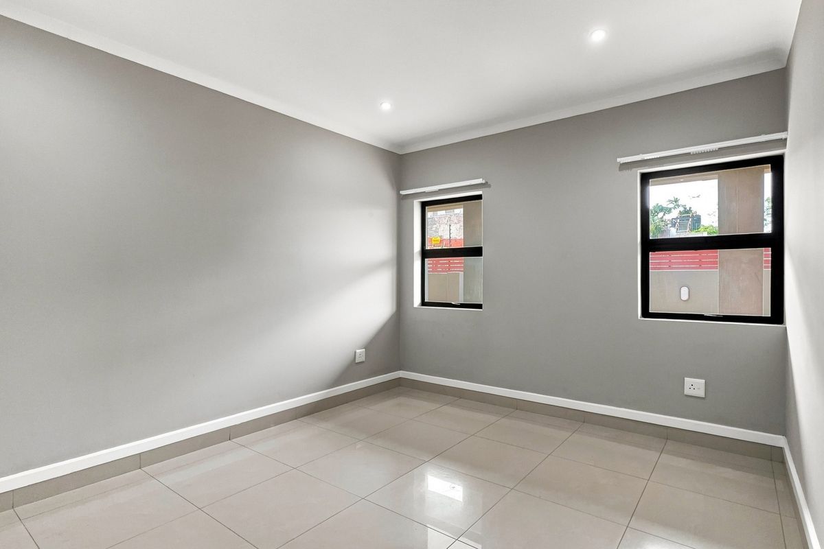 A bright, empty bedroom featuring neutral grey walls, large aluminium-framed windows, and modern tiled flooring with recessed ceiling lighting.