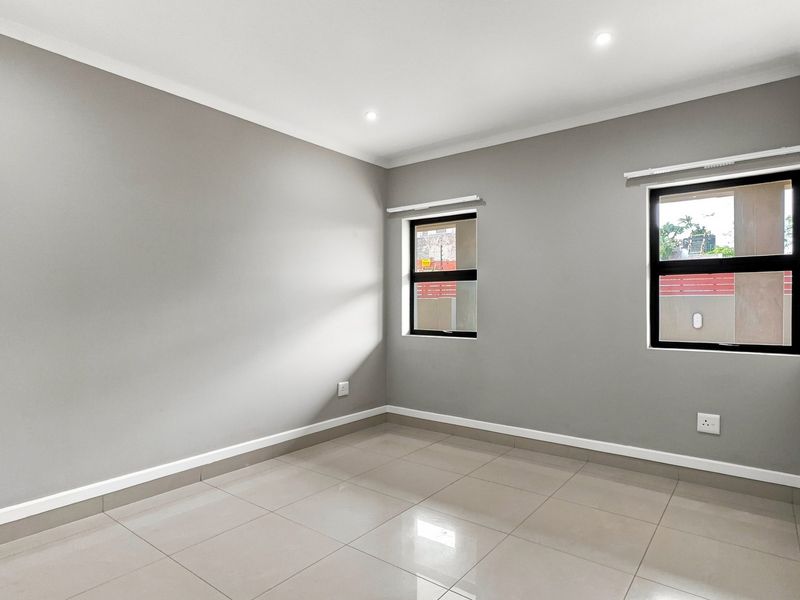 A bright, empty bedroom featuring neutral grey walls, large aluminium-framed windows, and modern tiled flooring with recessed ceiling lighting.