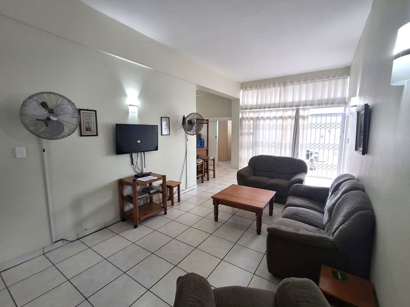 A spacious living room featuring neutral tiled flooring, wall-mounted lighting, and large windows with curtains providing access to the exterior through a security gate.