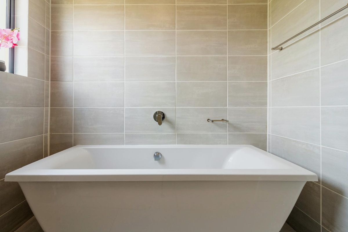 A clean, modern bathroom featuring a white bathtub with wall-mounted mixer tap and neutral ceramic wall tiling.