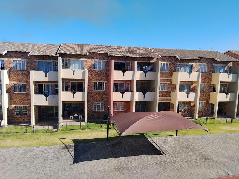 Exterior view of a multi-story apartment complex featuring face brick construction, private balconies, and a paved parking area with shade cloth carports.