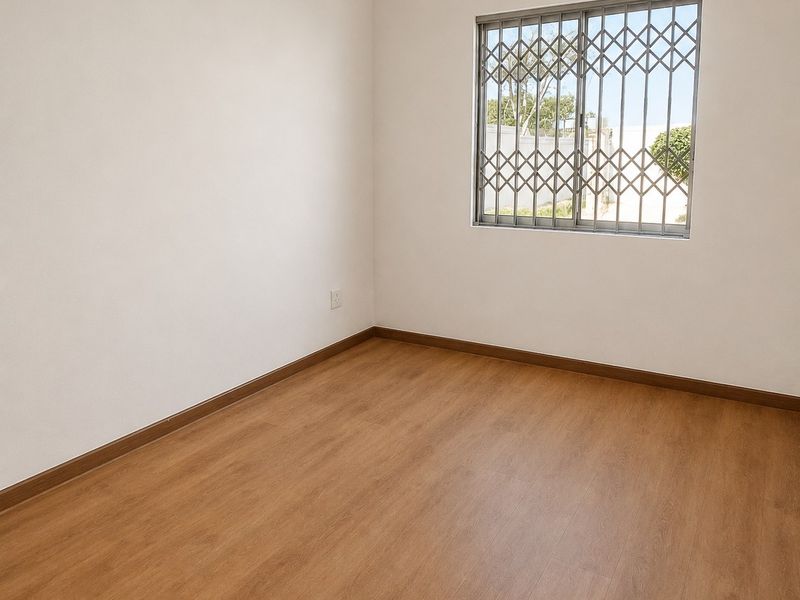 A clean, empty bedroom featuring light wood-look laminate flooring, neutral walls, and a window fitted with a security gate.
