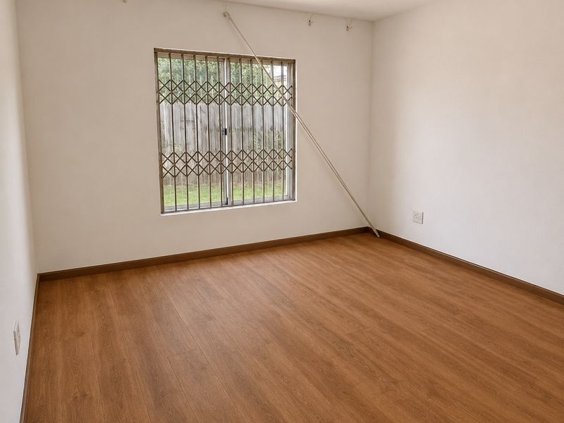 A bright, empty bedroom featuring wood-look laminate flooring, neutral walls, and a window fitted with security bars and a security gate.