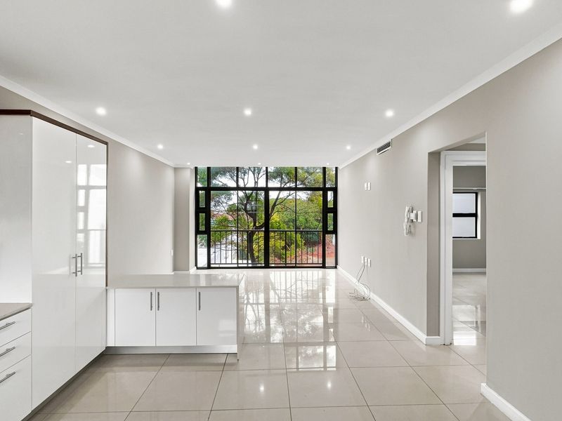 Modern open-plan living area featuring sleek white cabinetry, polished porcelain tiles, and large windows providing natural light and a pleasant leafy outlook.