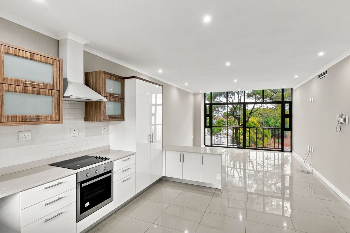 Modern open-plan kitchen and living area featuring sleek white and wood-grain cabinetry, stone countertops, integrated appliances, and large windows with a leafy outlook.