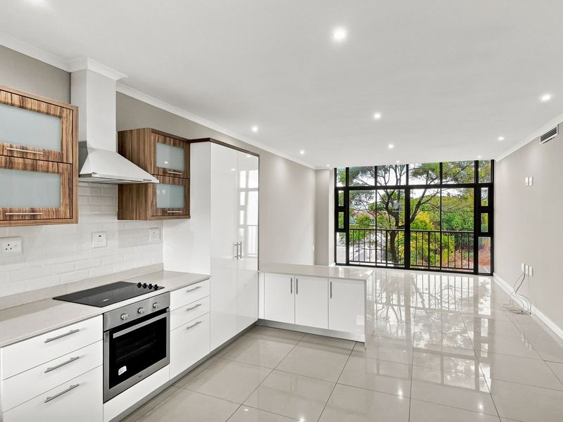 Modern open-plan kitchen and living area featuring sleek white and wood-grain cabinetry, stone countertops, integrated appliances, and large windows with a leafy outlook.