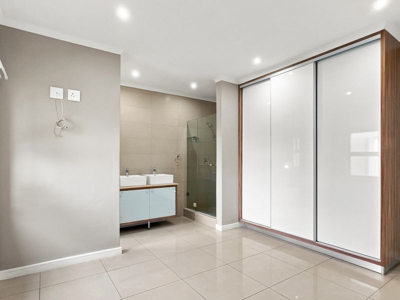 Spacious bedroom featuring modern built-in cupboards, a sleek double vanity, and an en-suite glass shower enclosure with neutral tiled flooring.