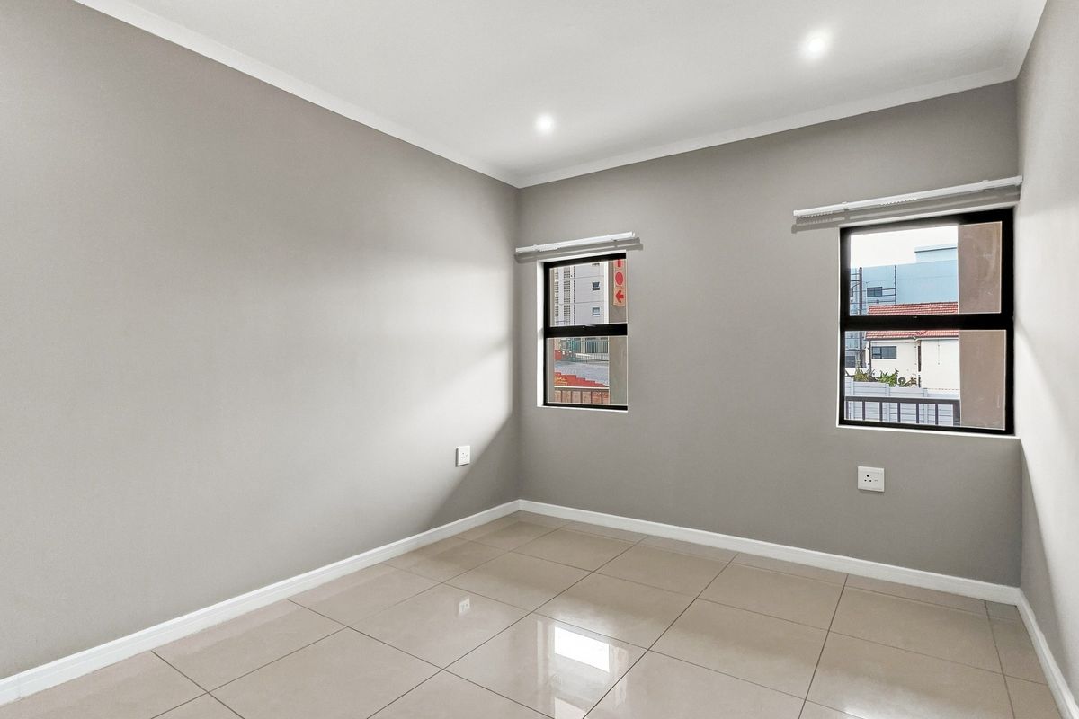 A bright, empty bedroom featuring neutral grey walls, large aluminium-framed windows, and polished floor tiles, offering a clean and versatile living space.