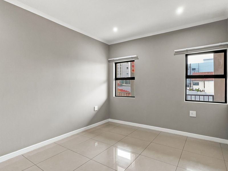 A bright, empty bedroom featuring neutral grey walls, large aluminium-framed windows, and polished floor tiles, offering a clean and versatile living space.