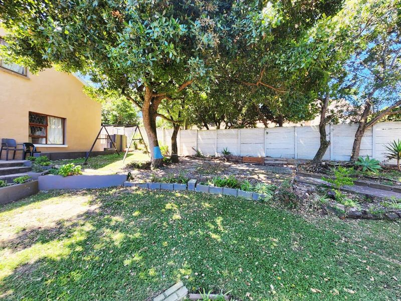 A rear garden area featuring a lawn, mature trees, a timber boundary fence, and a paved path with concrete retaining walls.