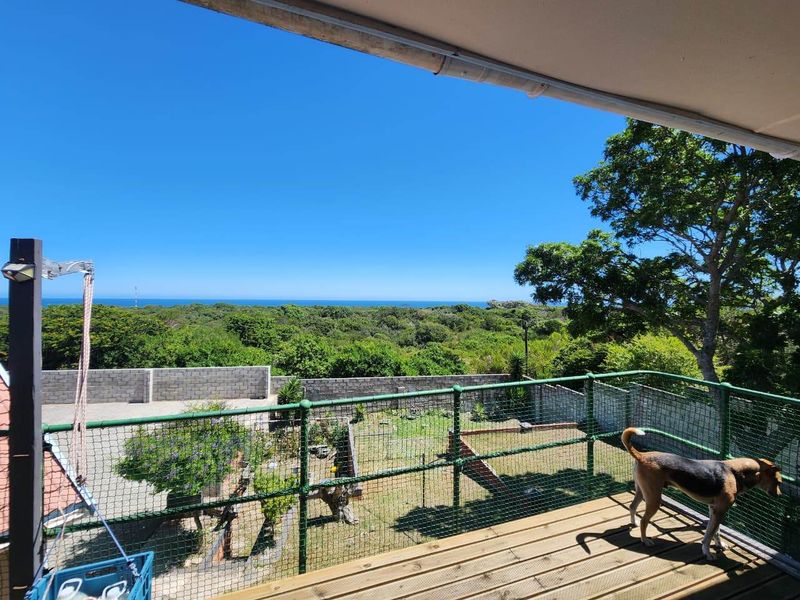 An elevated wooden balcony featuring a green metal mesh railing, offering expansive views over the garden and surrounding coastal landscape.