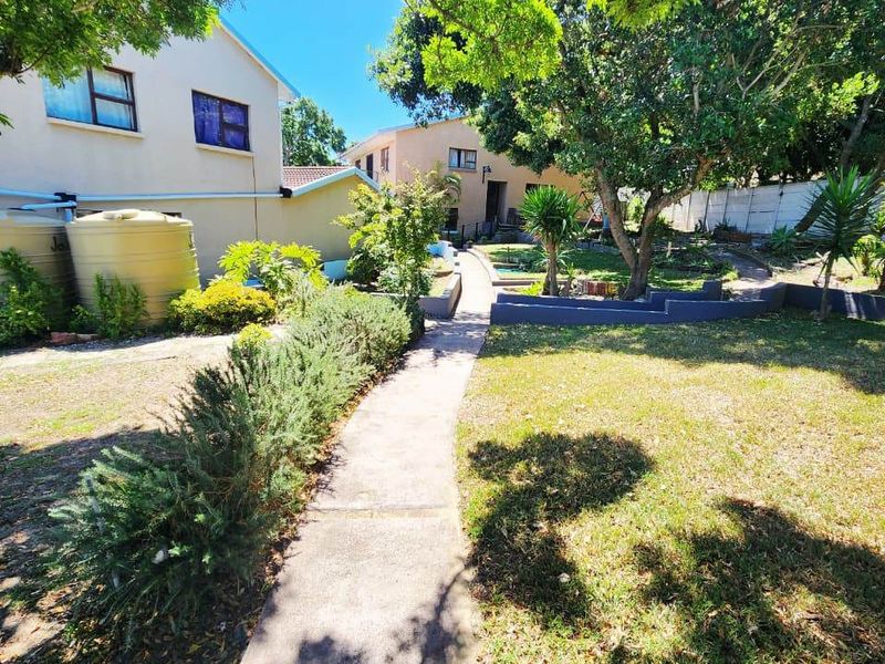 A spacious rear garden featuring a concrete pathway, mature trees, established landscaping, and water storage tanks alongside the property exterior.