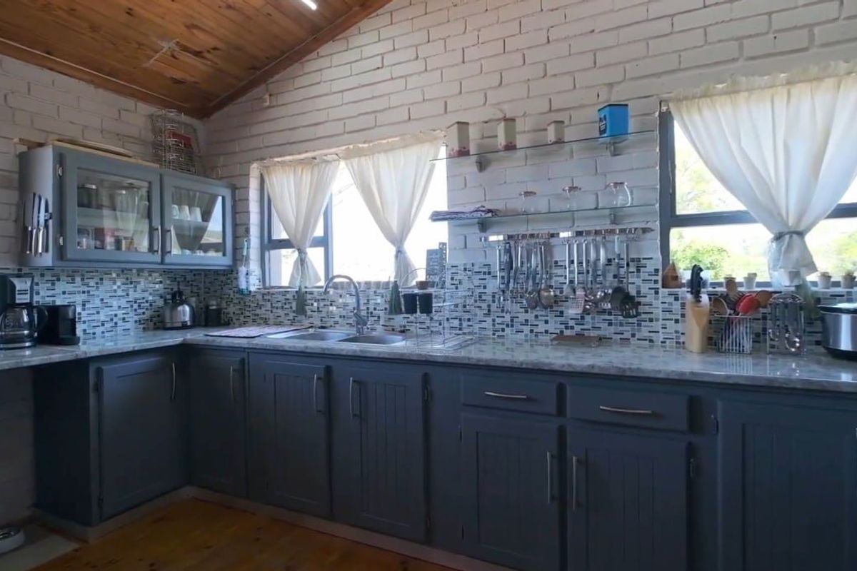 A well-lit kitchen featuring grey cabinetry, stone-look countertops, mosaic tile backsplash, and exposed timber ceilings with painted brick walls.