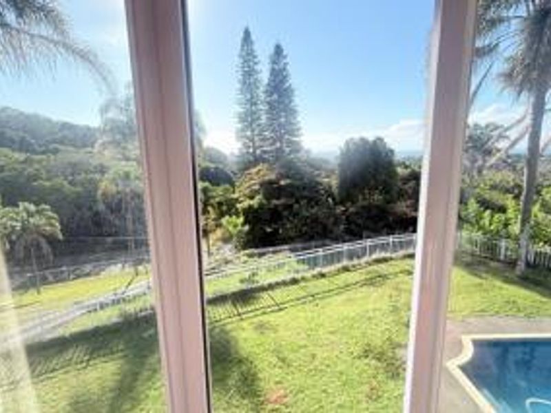 A scenic elevated view overlooking a lush green garden, swimming pool, and surrounding landscape, captured through a window.