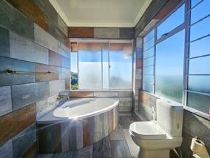 A bright bathroom featuring a corner bathtub, modern multi-toned tiled walls, and large windows with security bars offering natural light.