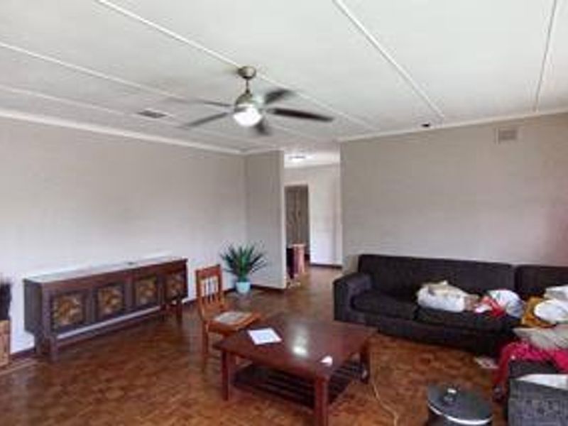 A spacious living room featuring classic parquet flooring, a central ceiling fan, and neutral wall finishes, offering a versatile layout for comfortable family living.
