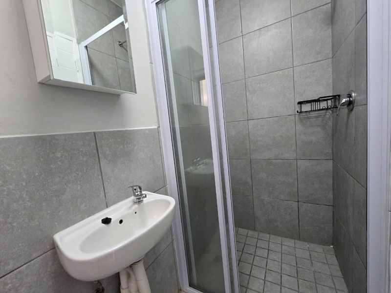A compact bathroom featuring a wall-mounted basin, a tiled shower enclosure, and a mirrored medicine cabinet with neutral grey wall tiling.