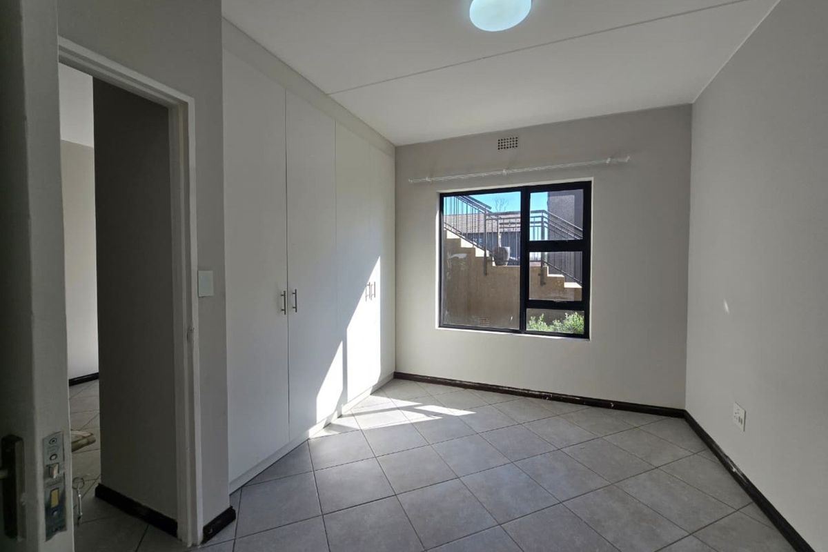 A clean, empty bedroom featuring neutral tiled flooring, built-in cupboards, and an aluminium-framed window providing natural light.