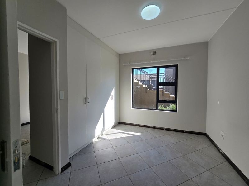 A clean, empty bedroom featuring neutral tiled flooring, built-in cupboards, and an aluminium-framed window providing natural light.