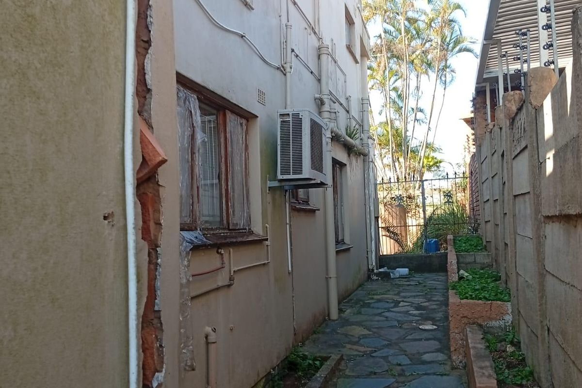 Side passage of the property featuring a stone paved walkway, air conditioning unit, and boundary wall with electric fencing, showing significant exterior wall damage.