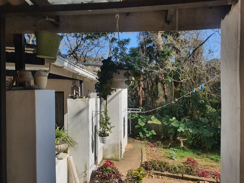 A covered patio area overlooking a garden, featuring concrete pathways and exterior wall access, requiring some maintenance and cosmetic updates.