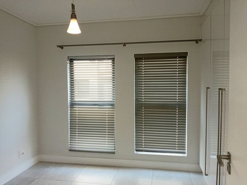 A bright, empty bedroom featuring tiled flooring, built-in cupboards, and dual windows fitted with venetian blinds.