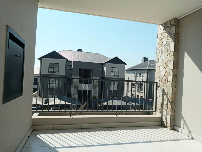 A clean, modern covered balcony featuring tiled flooring, a metal balustrade, and a decorative stone-clad pillar, offering an open view of the complex.
