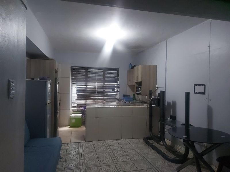 Open plan living and kitchen area featuring tiled flooring, built-in cabinetry, and window blinds. The space shows signs of ceiling water damage and exposed wiring.