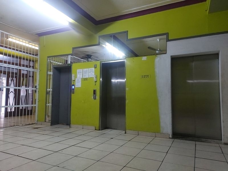 Apartment building lobby area featuring multiple elevators, security gate access, and tiled flooring, currently showing signs of wear and maintenance needs.