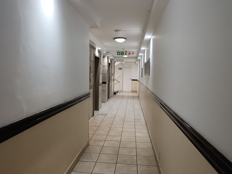 A functional apartment building hallway featuring tiled flooring, elevator access, and emergency exit signage with neutral wall finishes.