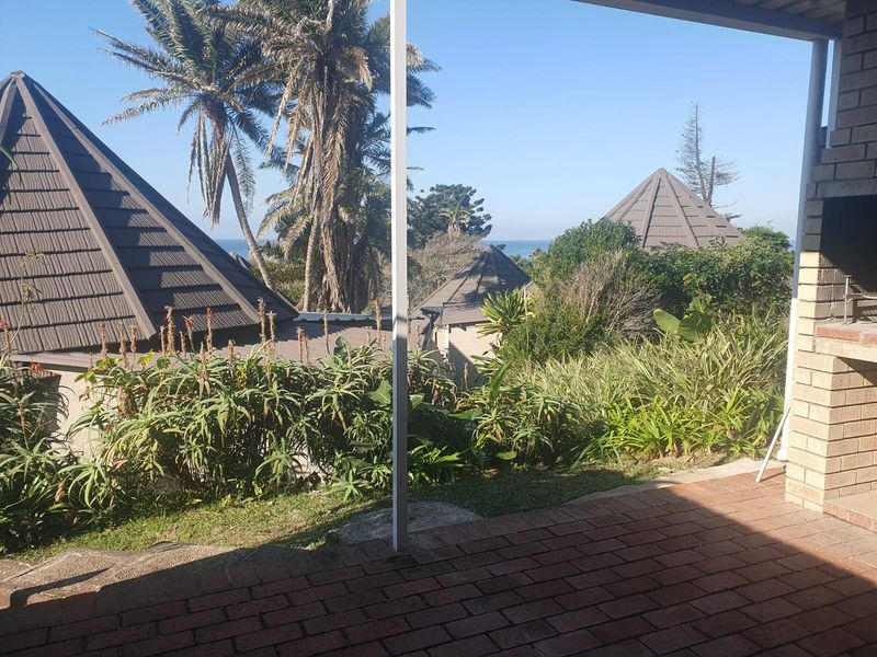 A covered outdoor patio featuring a brick built-in braai and paved flooring, overlooking a garden with distant sea views.