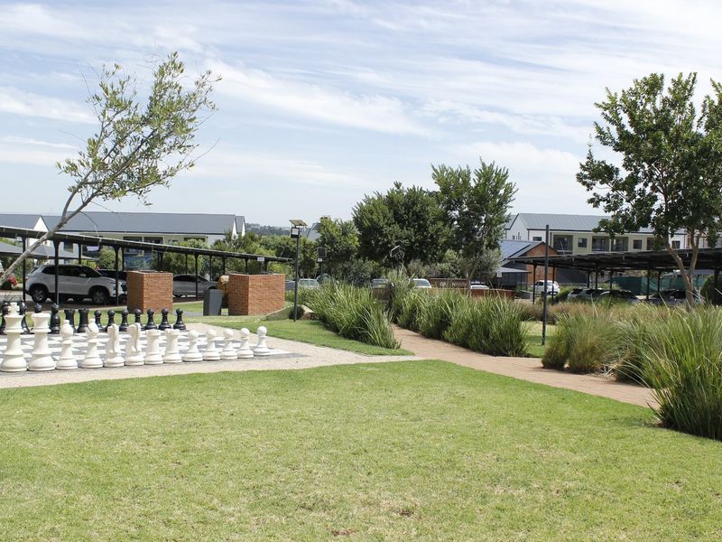 A well-maintained communal garden area featuring a paved walkway, landscaped lawn, and an outdoor chess set within an apartment complex.