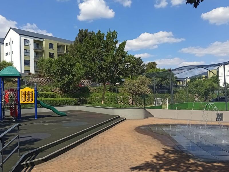 Communal outdoor recreational area featuring a playground, water feature, and fenced sports court within the apartment complex grounds.