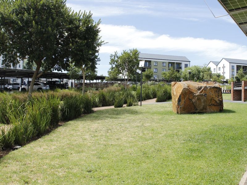 A well-maintained communal garden area featuring a paved walkway, brick braai facility, and covered parking structures within an apartment complex.