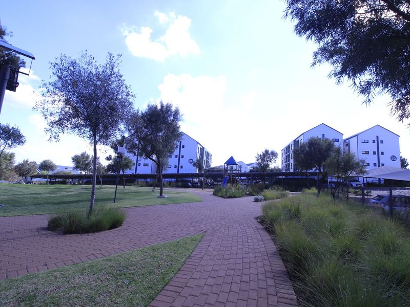 A well-maintained communal garden area featuring paved walkways, landscaped lawns, and mature trees within an apartment complex setting.
