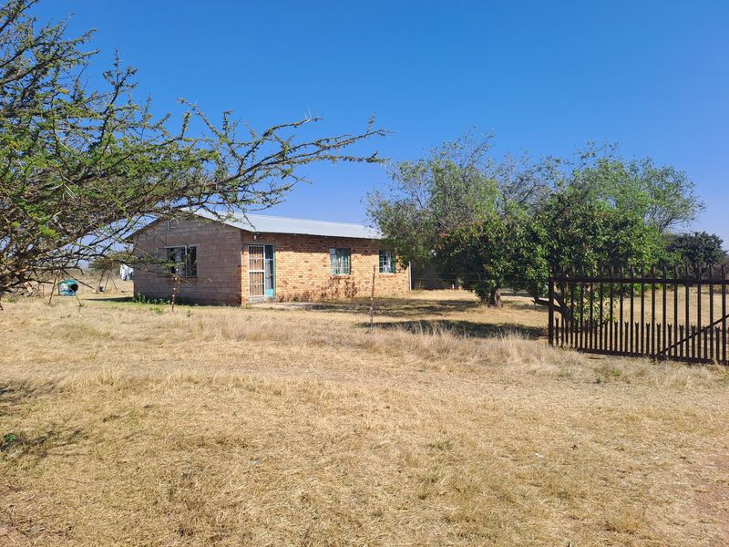 A single-story brick farmhouse exterior featuring a corrugated metal roof and secure metal perimeter fencing set on a rural property.