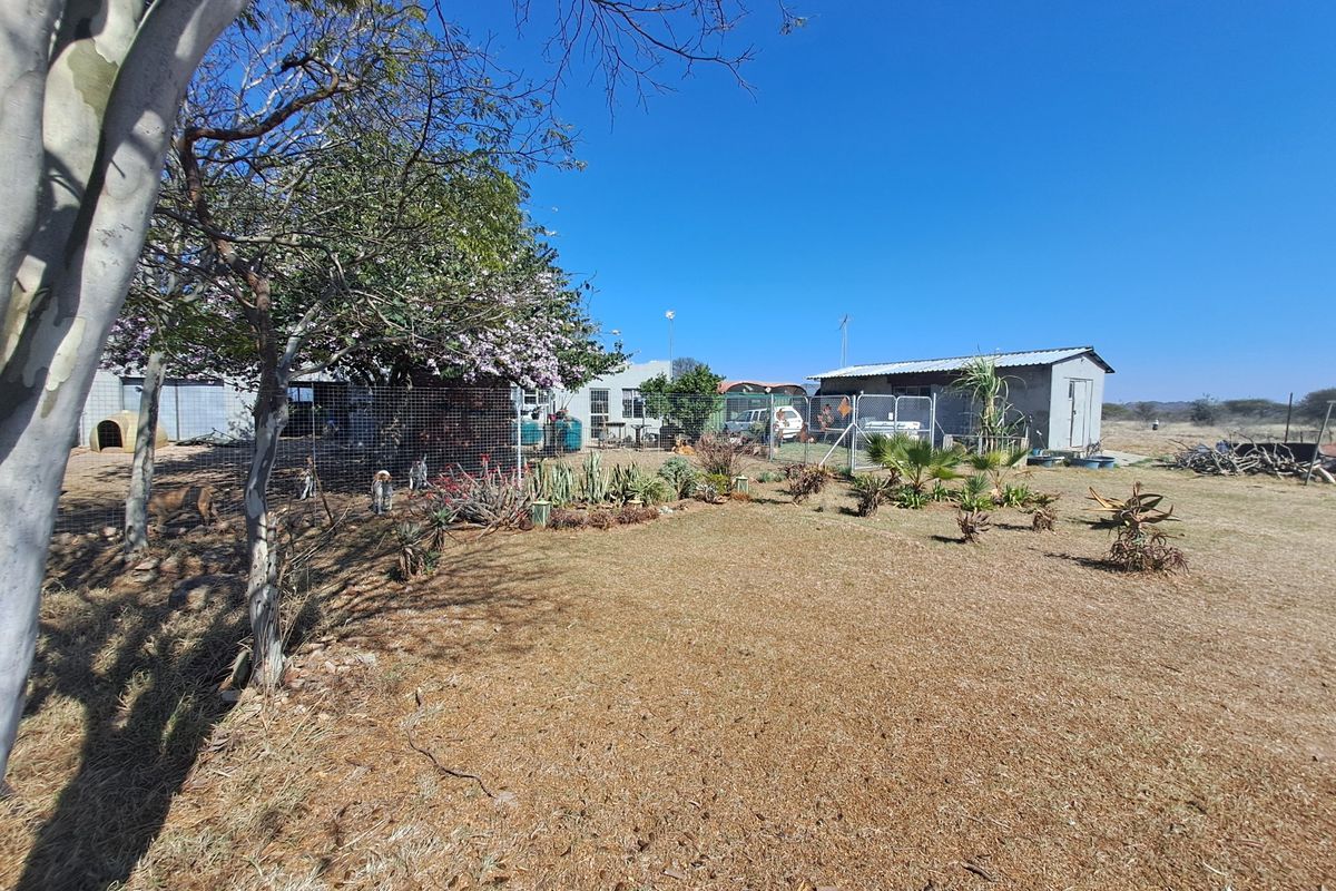 A spacious farm garden area featuring dry grass, mature trees, and functional wire mesh fencing surrounding outbuildings and storage structures.