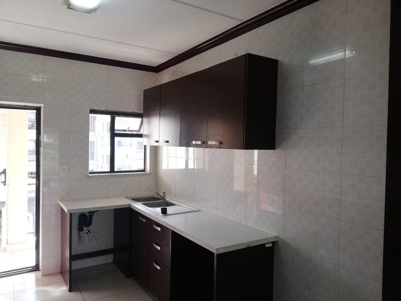A clean, empty kitchen featuring dark wood-finish cabinetry, white laminate countertops, a stainless steel sink, and full-height wall tiling.