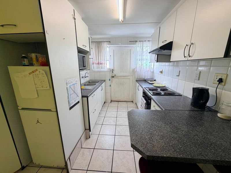 A functional galley-style kitchen featuring white cabinetry, dark laminate countertops, tiled flooring, and a built-in electric hob with an extractor fan.