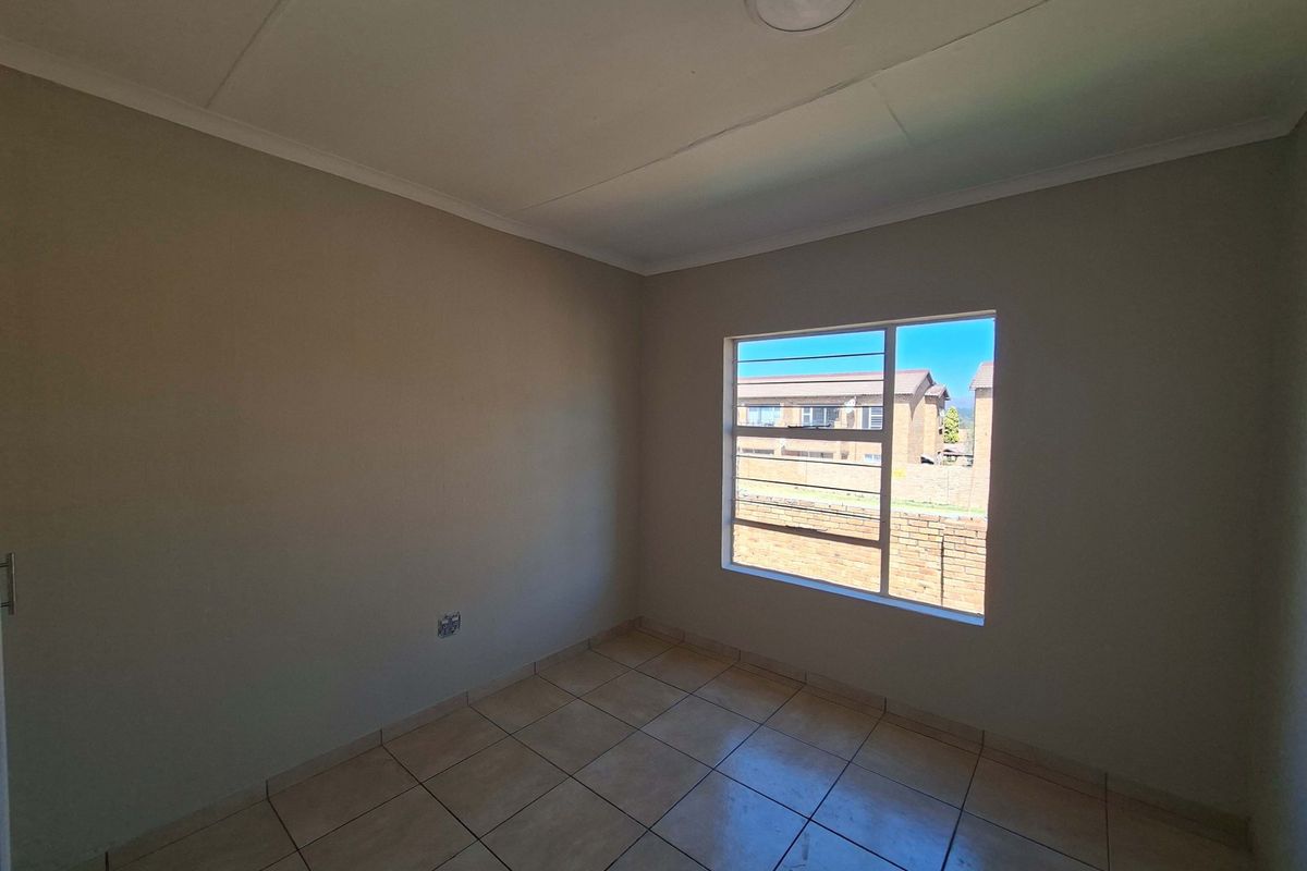A clean, empty bedroom featuring neutral wall tones, tiled flooring, and a large window providing natural light and a view of the surrounding complex.